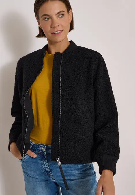 Vrouw in een zwarte jas, mosterdkleurige trui en blauwe jeans voor een neutrale achtergrond.