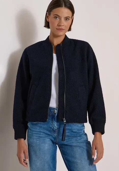 Vrouw met donkerblauw fleece bomberjack, wit T-shirt en blauwe spijkerbroek voor een neutrale achtergrond.