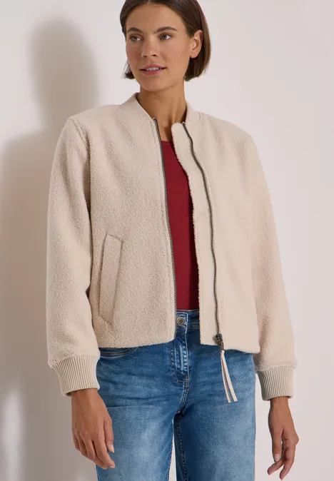 Frau in beiger Teddyjacke, rotem Shirt und blauen Jeans, stehend vor neutralem Hintergrund.