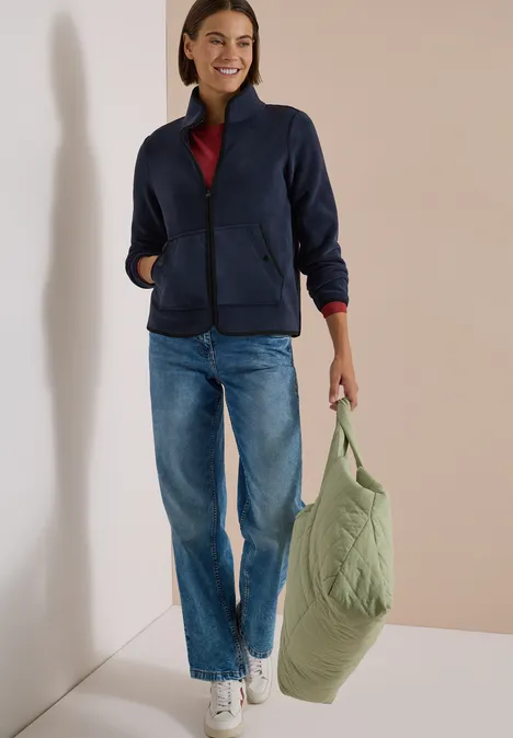 Frau in Jeans, marineblauer Jacke und rotem Shirt trägt eine grüne Stepp-Tasche.