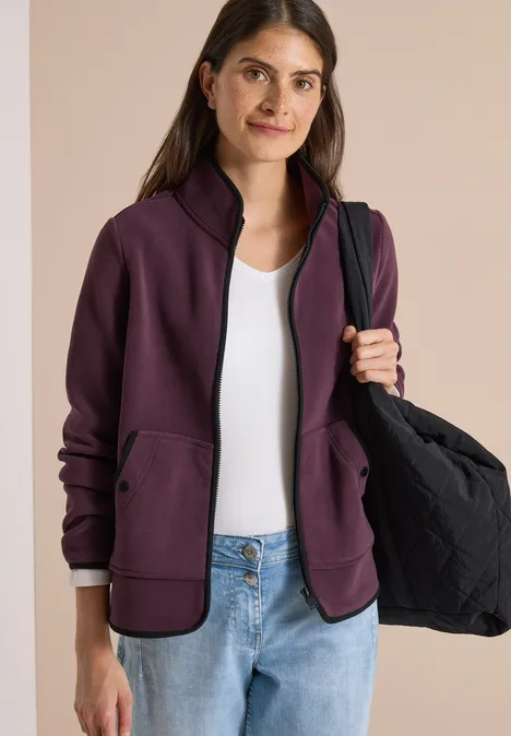 Vrouw draagt paarse jas, wit topje, spijkerbroek en houdt een zwarte tas.