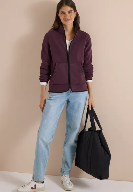 Een jonge vrouw draagt een paarse jas, wit shirt, lichtblauwe jeans, witte sneakers en een zwarte tas.