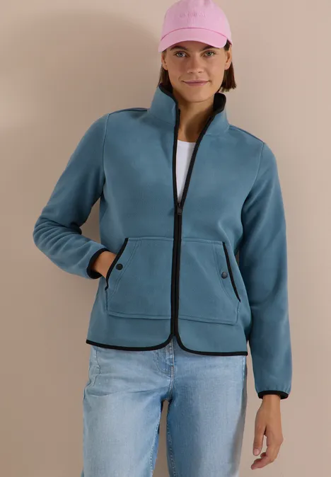 Frau trägt petrolfarbene Fleecejacke mit Reißverschluss, Taschen, über weißem Shirt, rosa Cap, Jeans.