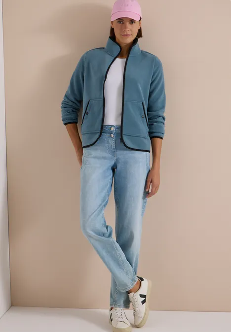 Eine Frau steht in Jeans, weißem Shirt, blauem Fleece, rosa Cap und Sneakern vor beige.