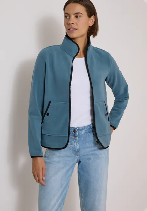 Frau trägt türkisblaue Fleecejacke mit Reißverschluss und schwarzen Details, dazu weißes T-Shirt und Jeans.