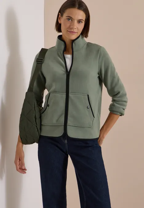 Frau trägt grüne Fleecejacke, weißes Shirt, blaue Jeans und grüne Umhängetasche vor hellem Hintergrund.