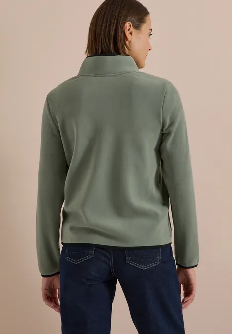 Person von hinten in grünem Fleecepulli und dunkler Jeans vor beigem Hintergrund.