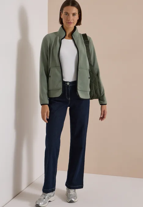 Frau steht in Raumecke, trägt dunkle Jeans, weiße Bluse, grüne Jacke und Sneaker.