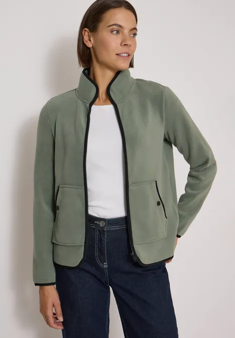 Frau trägt olivgrüne Fleecejacke mit Reißverschluss und Taschen, kombiniert mit weißem Shirt und Jeans.