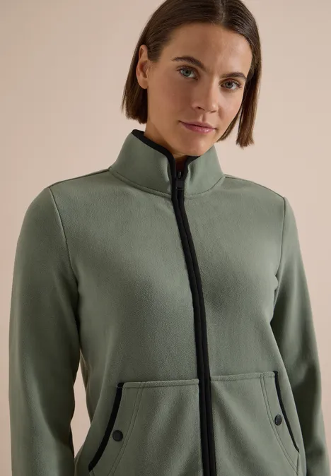 Frau trägt olivegrüne Fleecejacke mit Stehkragen und Reißverschluss, Kängurutaschen, schwarze Details