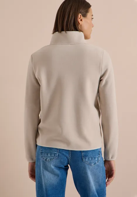 Eine Frau steht mit dem Rücken zur Kamera und trägt einen hellbeigen Fleecetop mit Stehkragen und blauen Jeans.