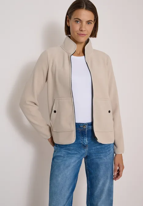 Frau trägt beige Jacke, weißes Shirt und blaue Jeans vor hellem Hintergrund.