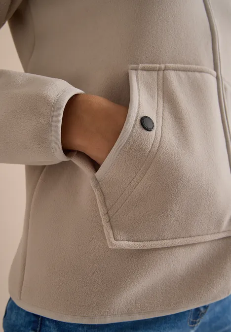 Beige Fleecejacke mit aufgesetzter Tasche und Druckknopf, Hand steckt in der Tasche.