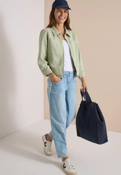 Frau im lässigen Look mit grüner Overshirt-Jacke, weißem Shirt, weiten Jeans, Sneakers, blauer Tasche und Cap.