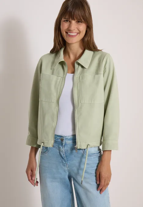 Eine lächelnde Frau in hellgrüner Jacke, weißem Top und Jeans vor neutralem Hintergrund.
