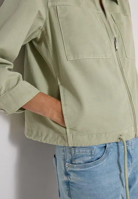 Person trägt eine helle, olivgrüne Jacke mit eingesteckter Hand in der Tasche und hellblaue Jeans.