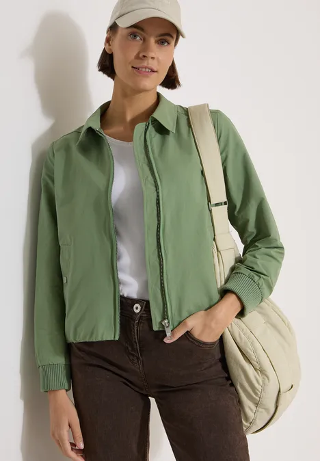 Blouson à col chemise deep matcha green Une personne porte une veste verte, un t-shirt blanc, un pantalon marron et un sac beige.