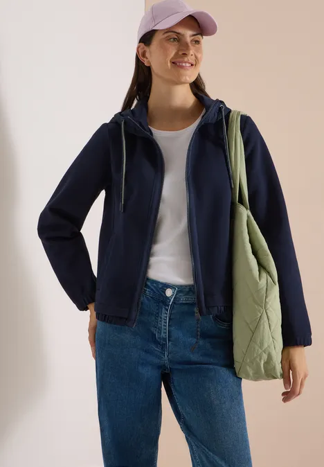 Vrouw in donkerblauwe hoodie, wit T-shirt, spijkerbroek en groene gewatteerde tas met lila pet, glimlachend