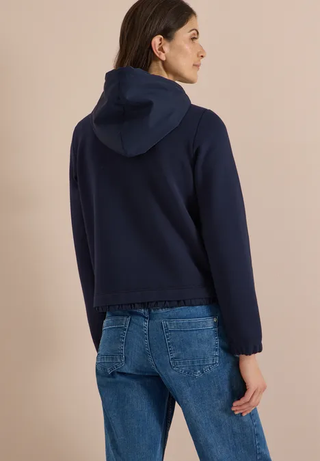 Vrouw van achteren voor een beige achtergrond, draagt een donkerblauwe capuchontrui en een jeans.
