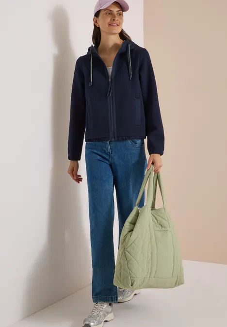 Een vrouw draagt een donkerblauwe hoodie, jeans, een pet en houdt een grote lichtgroene, gewatteerde tas vast.