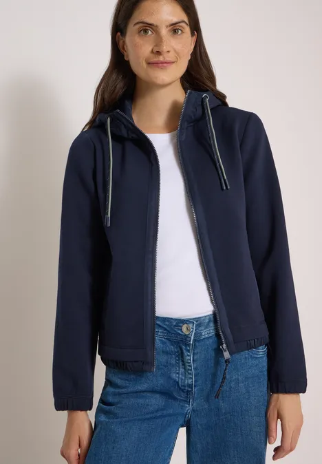 Vrouw in donkerblauwe capuchontrui over wit shirt en blauwe spijkerbroek voor een lichte achtergrond.