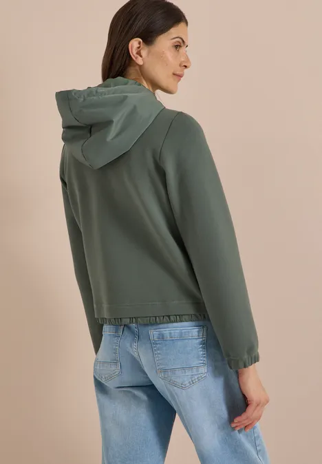Eine Frau steht seitlich von hinten, trägt eine grüne Kapuzenjacke und blaue Jeans.