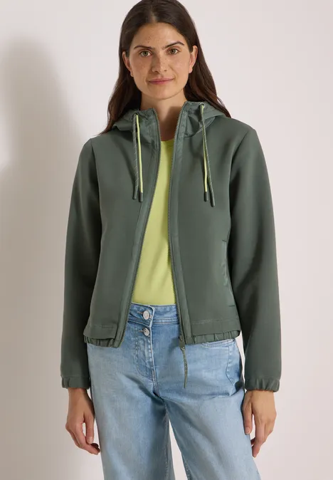 Frau in grüner Kapuzenjacke, gelbem Shirt und Jeans steht vor hellem Hintergrund.