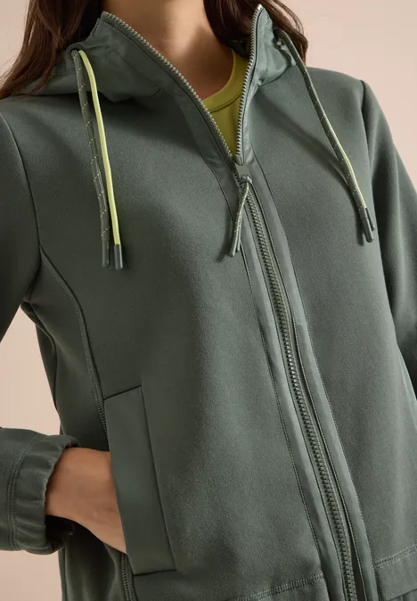 Person trägt graugrünen Kapuzenpullover mit Reißverschluss, seitlichen Taschen und hellgrünen Kordeln.