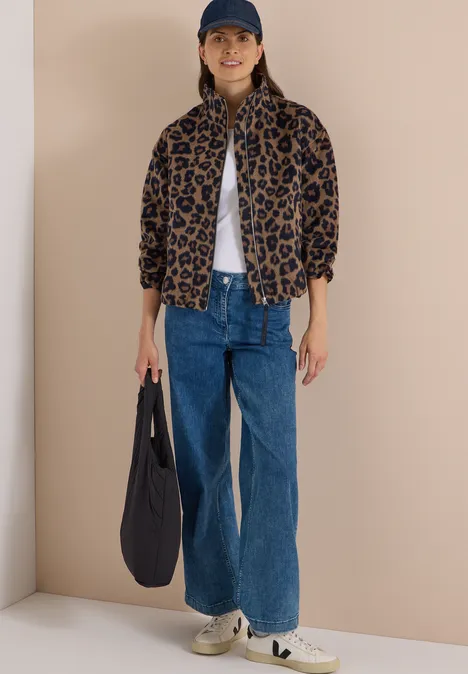 Vrouw draagt een luipaardprintjas, wit T-shirt, blauwe jeans, sneakers, pet en draagt een donkerblauwe tas.