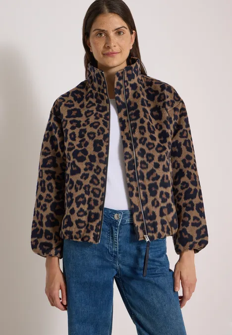 Vrouw in een korte jas met luipaardprint, rits en wijdvallende mouwen, gecombineerd met een blauwe spijkerbroek.