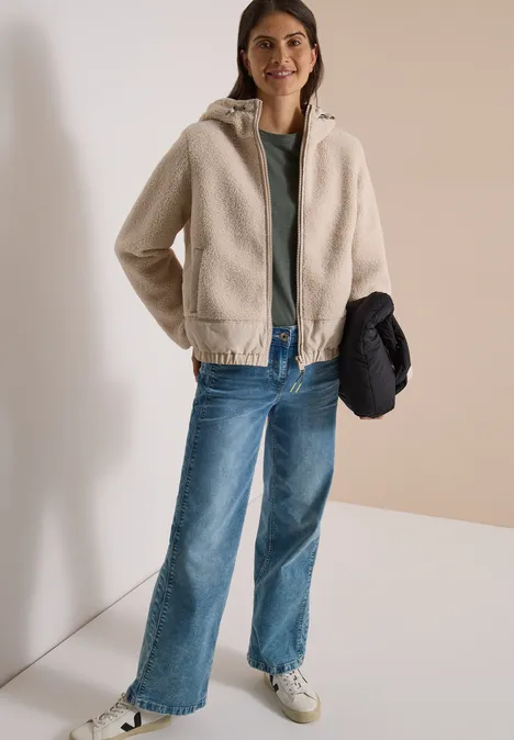 Teddy-Jacke mit Softshell-Details teddy vanilla white Frau in beiger Teddyjacke, grünem Shirt, blauen Jeans und weißen Sneakern, hält dunkle Jacke in der Hand.