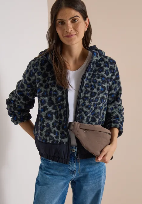 Vrouw met een gebloemde jas, wit shirt, spijkerbroek en een beige clutch lacht naar de camera