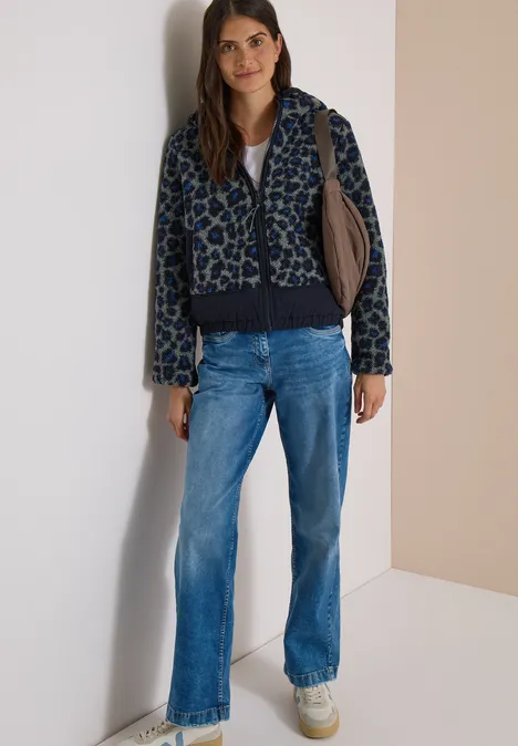 Vrouw in blauw-zwarte panterprint capuchonjack, rechte jeans en witte sneakers tegen lichte muur.