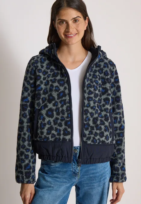 Een vrouw draagt een donkerblauwe capuchonvest met luipaardprint, een wit T-shirt en een blauwe jeans.