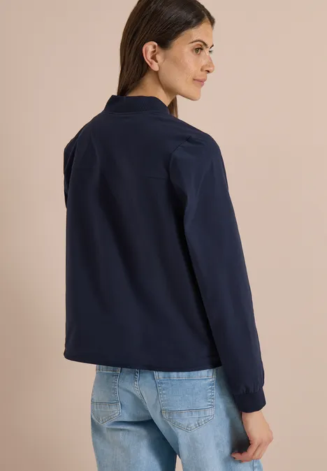 Vrouw van achteren in een donkerblauw shirt met lange mouwen en lichtblauwe jeans, voor een neutrale achtergrond.