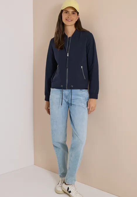 Een vrouw staat bij een muur, ze draagt een donkerblauw jasje, lichte jeans, witte sneakers en een gele pet.
