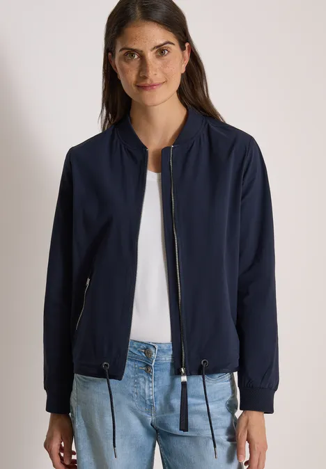 Vrouw draagt marineblauwe bomberjas, wit shirt en blauwe jeans.