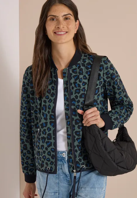 Frau trägt grüne, schwarz gepunktete Jacke über weißem Shirt, Jeans und schwarze gesteppte Tasche.