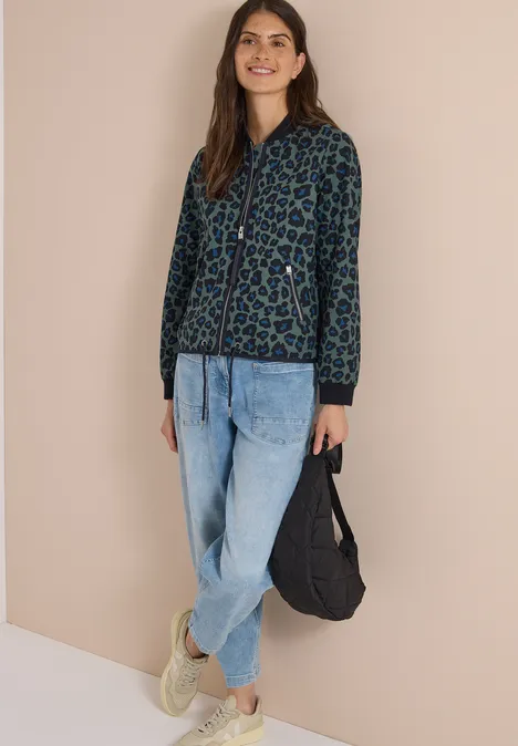 Frau trägt blau-grünen Leoprint-Cardigan, helle Jeans und Sneaker, hält schwarze Tasche und lächelt vor beiger Wand.