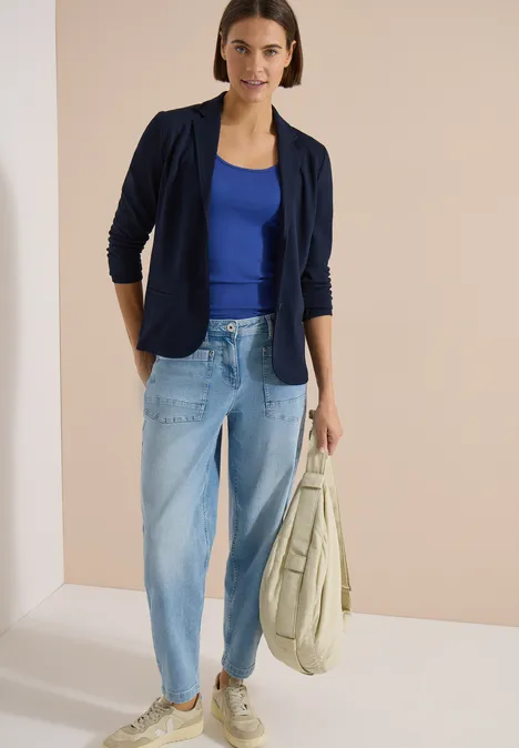 Frau trägt dunkelblauen Blazer, blaues Top, helle Jeans und hält eine große Tasche vor hellem Hintergrund.
