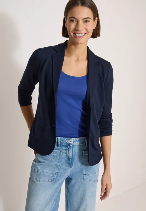 Frau mit dunkelblauem Blazer, blauem Top und hellblauen Jeans vor hellem Hintergrund.