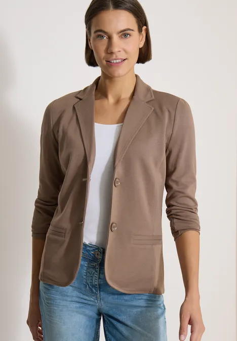 Frau in braunem Blazer, weißem Shirt und Jeans, stehend vor neutralem Hintergrund.
