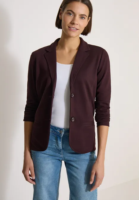 Frau im bordeauxroten Blazer mit weißem Top und blauen Jeans vor neutralem Hintergrund.