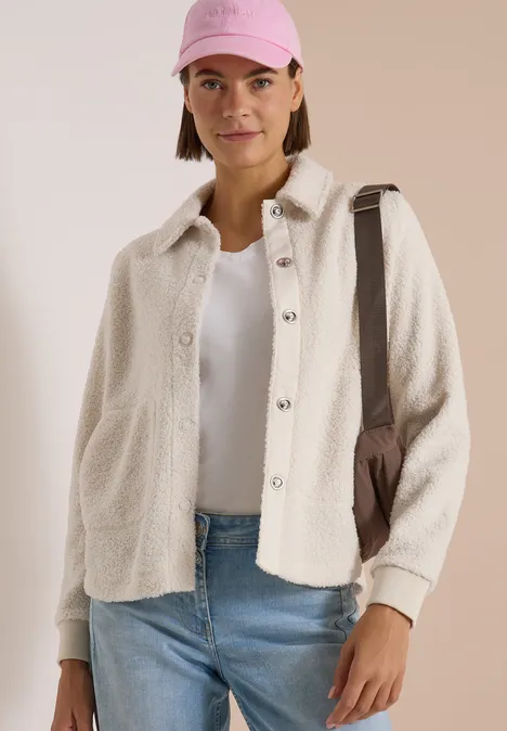 Frau mit rosa Cap, weißem T-Shirt, cremefarbener Hemdjacke, blauen Jeans und Umhängetasche.