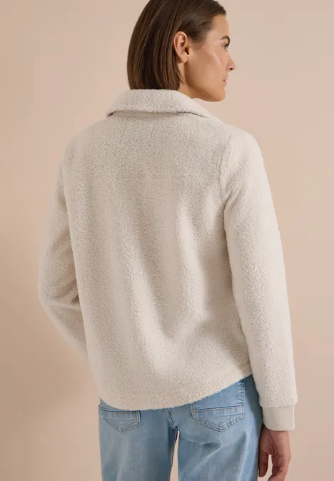 Frau von hinten trägt weiße Teddy-Fleece-Jacke und Jeans vor hellem Hintergrund.