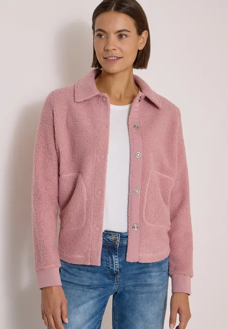 Eine Frau in rosa Fleecejacke, weißem Shirt und blauer Jeans steht vor hellem Hintergrund.