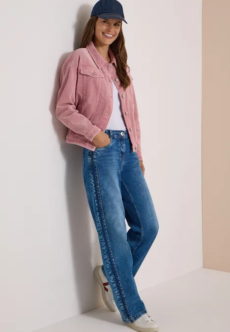 Une femme souriante en veste rose, jean et casquette, appuyée de façon décontractée contre un mur.