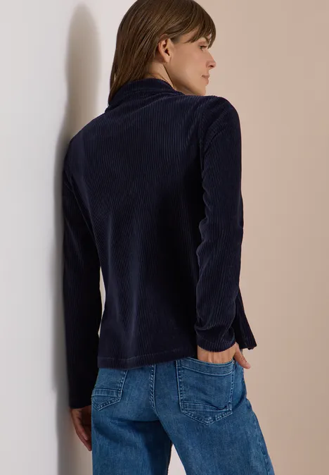 Frau im Profil, trägt dunkelblauen Samtblazer und Jeans, lehnt lässig an einer Wand.