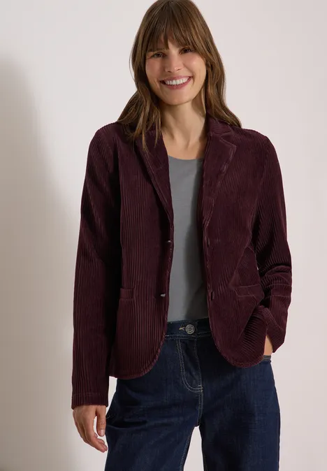 Eine lächelnde Frau trägt dunkelroten Cordblazer, graues Shirt und blaue Jeans.