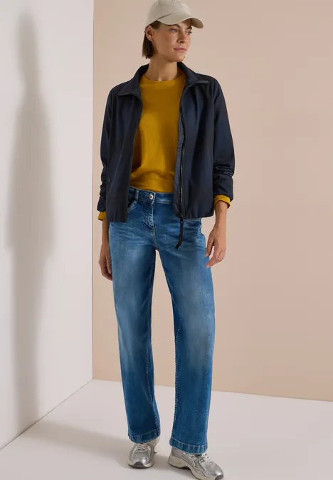 Frau trägt blaue Jeans, senfgelber Pullover, dunkelblaue Jacke und beige Mütze vor hellem Hintergrund.